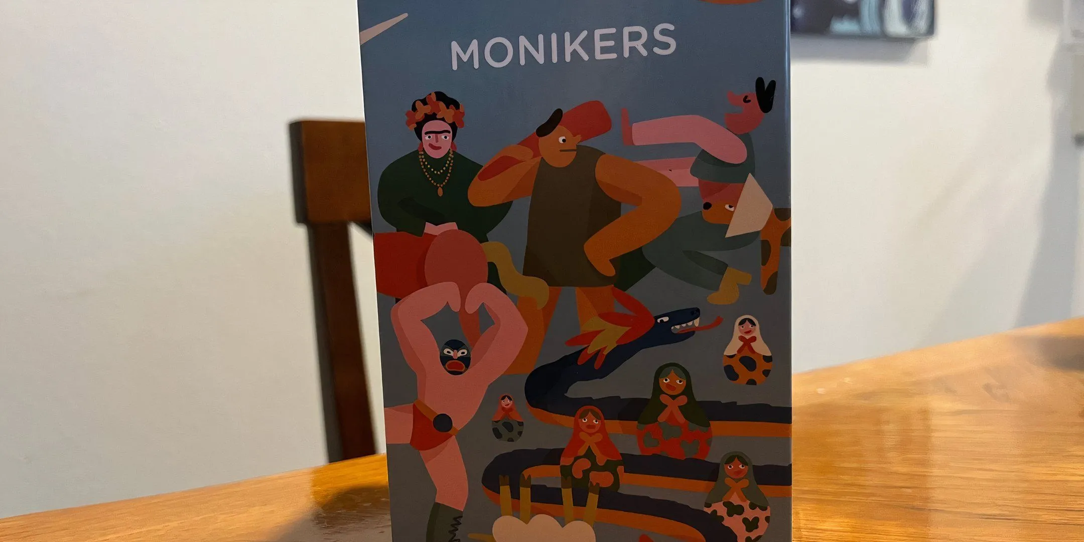 Bìa hộp game Monikers, gợi liên tưởng đến trò chơi đố từ ngữ và diễn kịch.